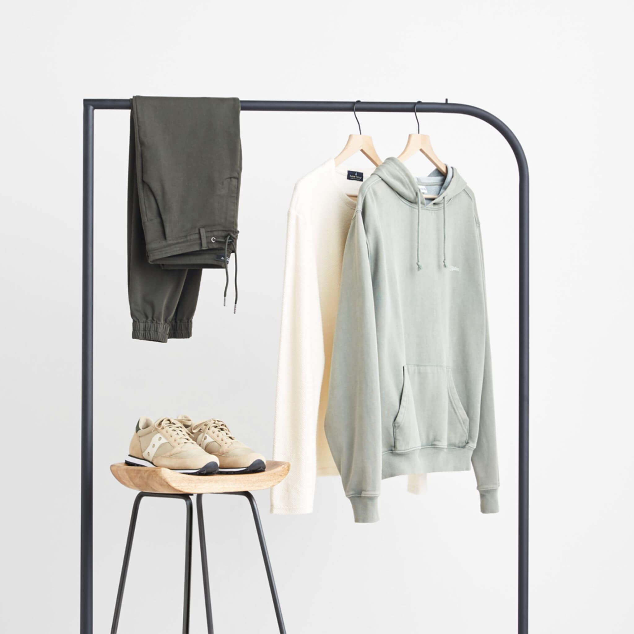 Stitch Fix Men’s grey hoodie, cream french terry pullover and dark grey woven joggers hanging on a black rack next to tan sneakers sitting on a wooden stool.